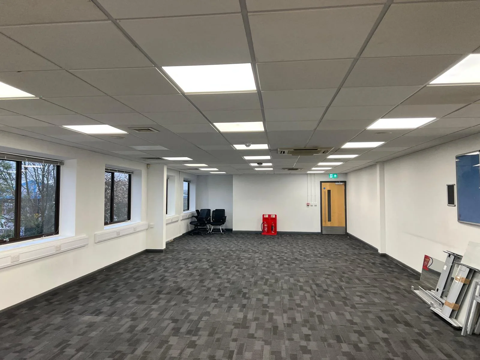 Wide view of a carpeted office with windows along the left wall and ceiling lights on.